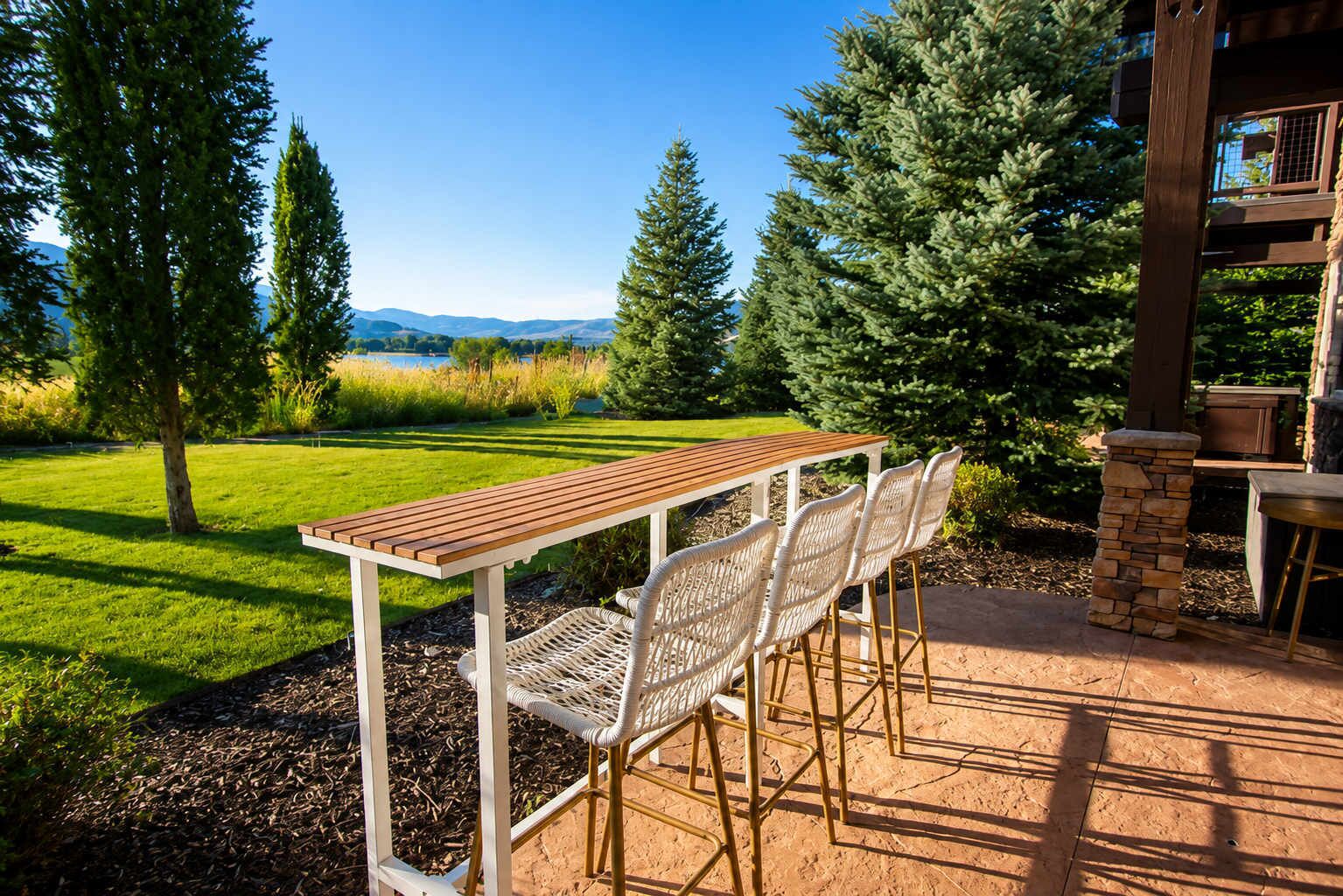 An outdoor patio with a wooden table and white wicker chairs, surrounded by a stone wall and a view of a lake and mountains.