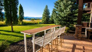 An outdoor patio with a wooden table and white wicker chairs, surrounded by a stone wall and a view of a lake and mountains.