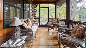 A cozy log cabin interior with wicker furniture, a ceiling fan, and large windows offering a view of the forest. - A Log Home Above the Lake | Hickory Hills Cabin (Highlands)