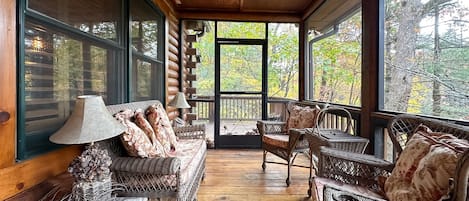 Interior acogedor de una cabaña de madera con muebles de mimbre, un ventilador de techo y grandes ventanas que ofrecen vistas al bosque.