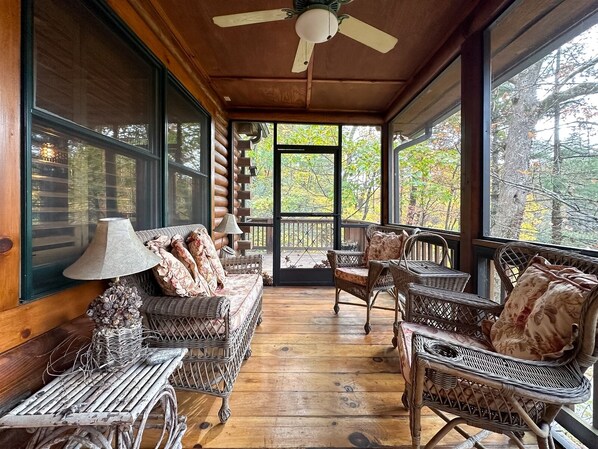 Interior acogedor de una cabaña de madera con muebles de mimbre, un ventilador de techo y grandes ventanas que ofrecen vistas al bosque.