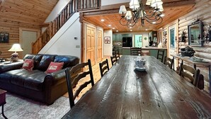A spacious dining area with a long wooden table and chairs, a black leather sofa, and a staircase leading to an upper level. - A Log Home Above the Lake | Hickory Hills Cabin (Highlands)