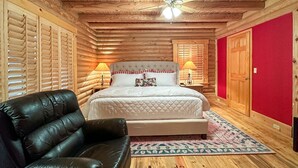 A log cabin bedroom with a bed, a leather chair, a ceiling fan, and a red wall. - A Log Home Above the Lake | Hickory Hills Cabin (Highlands)