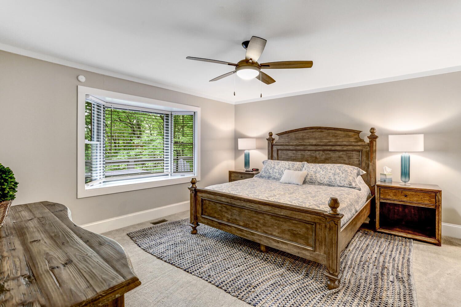 A bedroom with a wooden bed, a ceiling fan, a window with blinds, and a nightstand with a lamp.