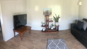 A living room with a flat-screen TV, a wooden coffee table, a black sofa, a grey rug, and a plant stand with decorative items. - Night retreat in the country (Paterson)