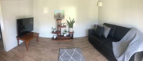 A living room with a flat-screen TV, a wooden coffee table, a black sofa, a grey rug, and a plant stand with decorative items.