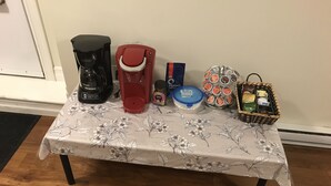 A table with a floral tablecloth, a black coffee maker, a red coffee maker, a basket of snacks, a container of Nespresso pods, and a basket of tea bags. - Stylish 1BR unit in Bedford, only 15 min to downtown Halifax. Near shopping area (Halifax)
