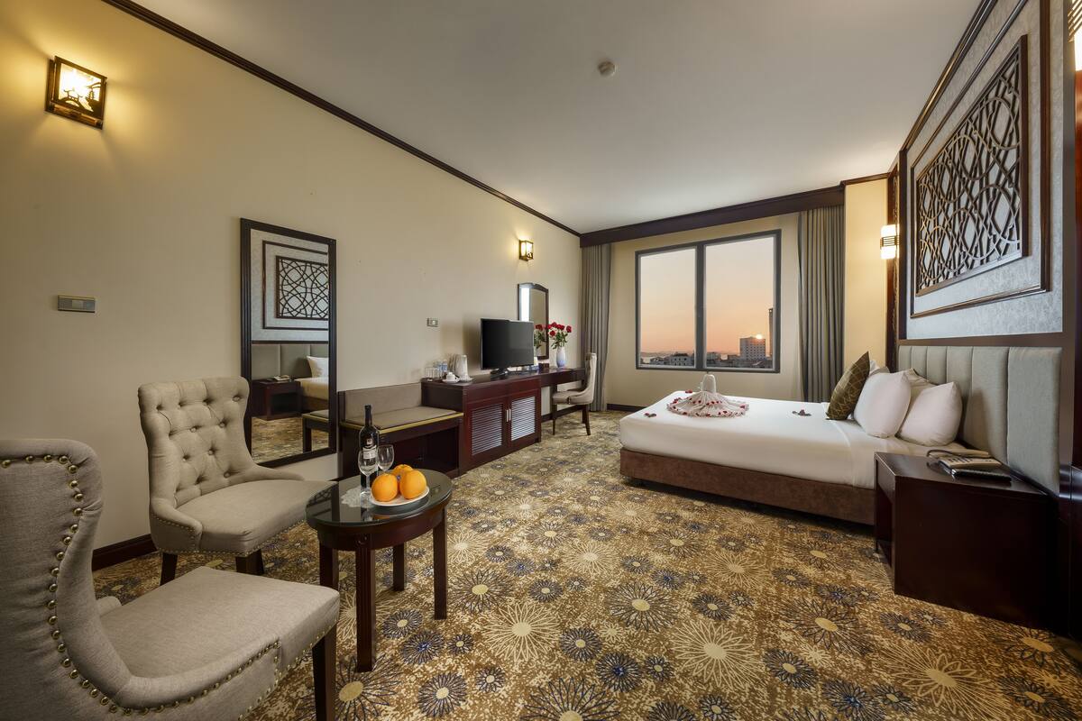 Habitación de hotel con cama, escritorio, silla, televisor y ventana grande con vistas a la ciudad.