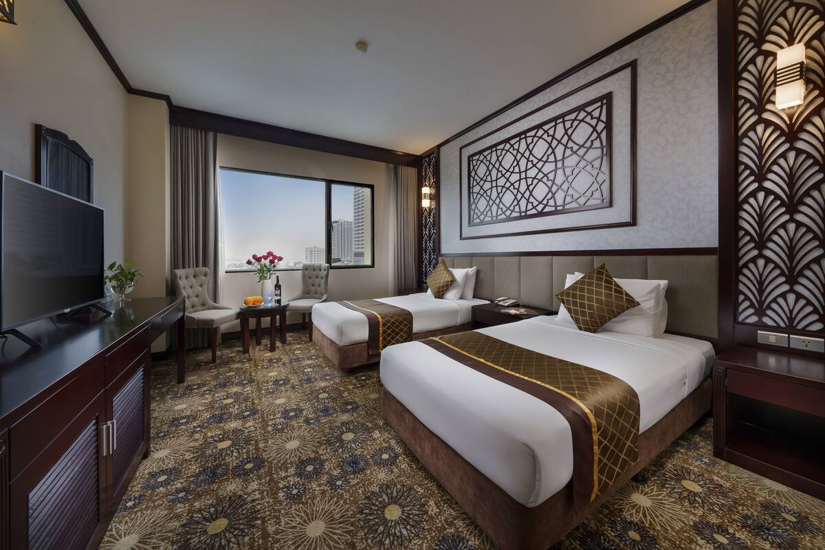 Habitación de hotel con dos camas, televisión, escritorio y ventana grande con vista a la ciudad.
