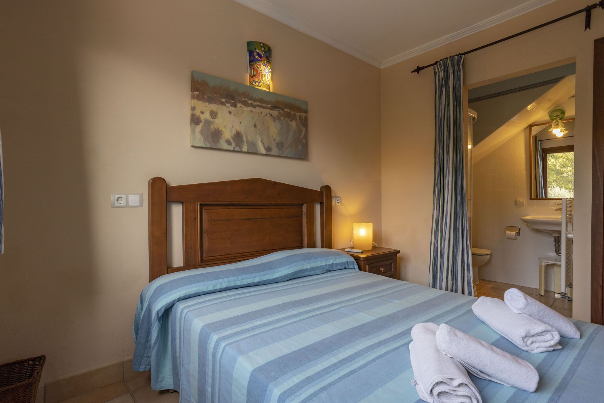 A bedroom with a bed, a nightstand, a lamp, a painting, and a bathroom visible through an open door.