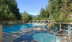 A pool area with lounge chairs and a wooden deck, surrounded by trees and mountains in the background. - Ski in/Ski out | Private hot tub | BBQ | Parking (Whistler)