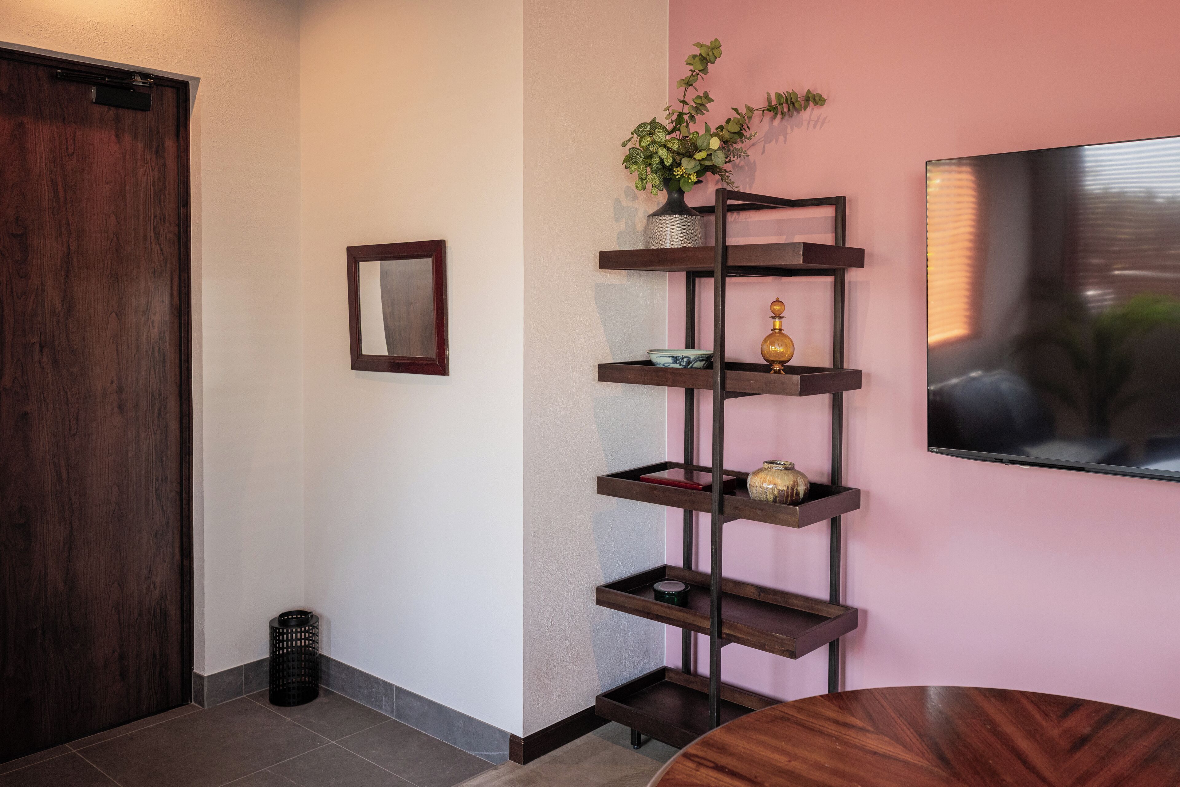 Un intérieur moderne avec une porte en bois, un téléviseur fixé au mur, une étagère à plusieurs niveaux ornée d’objets décoratifs et une table ronde en bois.