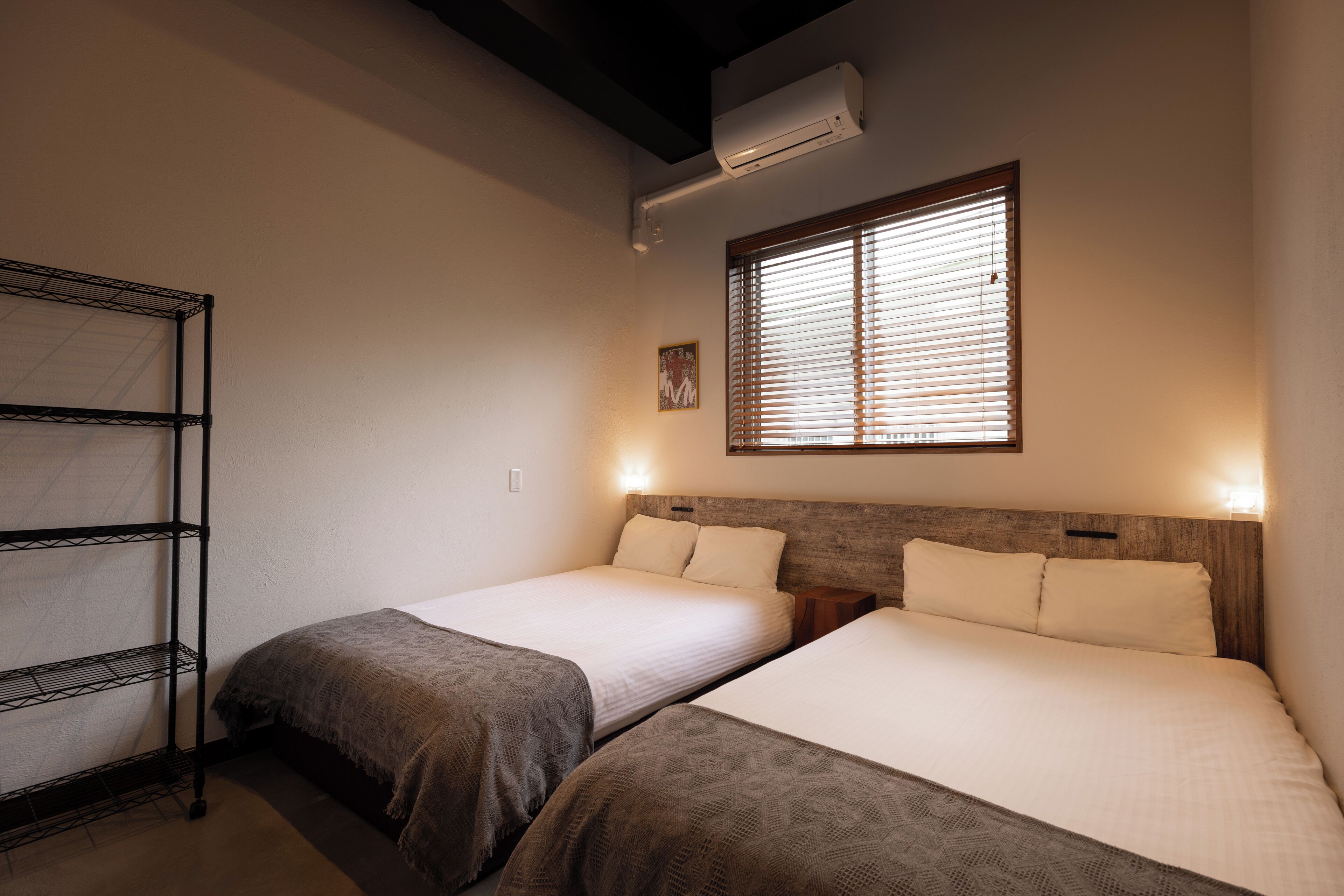 A hotel room with two beds, a metal shelf unit, and a window with blinds.