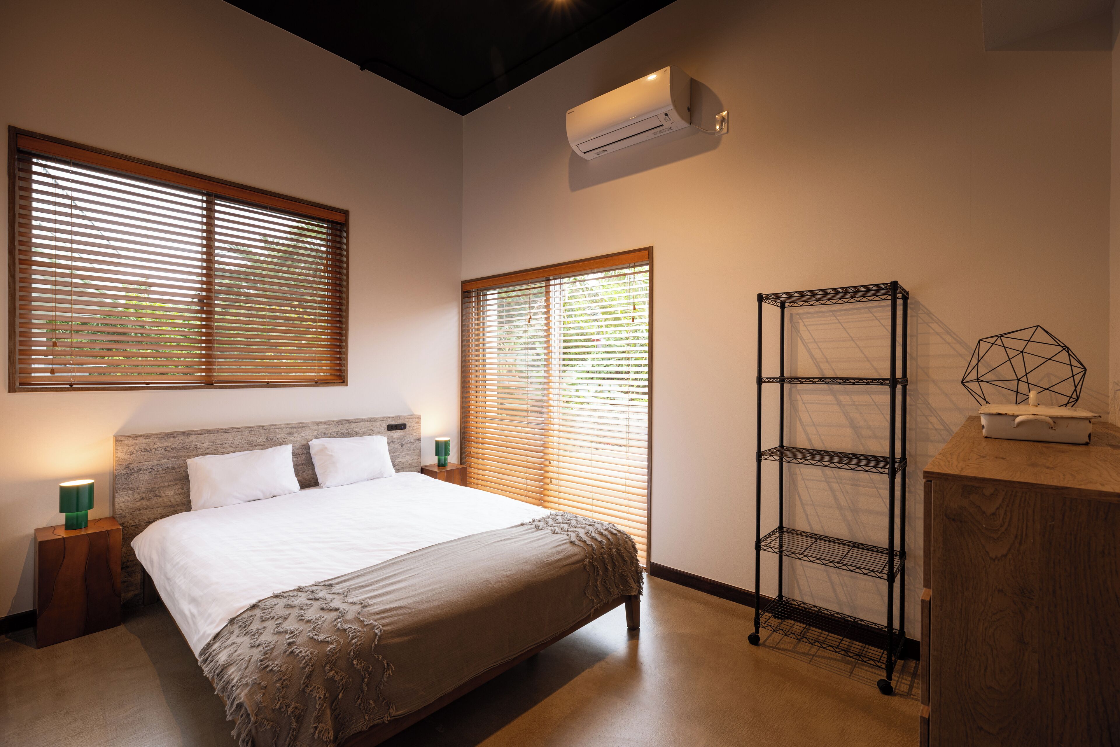 A bedroom with a bed, two bedside tables with lamps, a wall-mounted air conditioner, and a metal shelving unit.