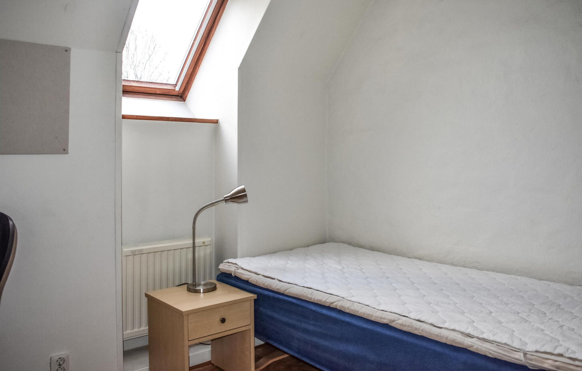 Een kleine kamer met een enkel bed, schuine kap, een radiator, een nachtkastje met lamp en een raam met houten kader.