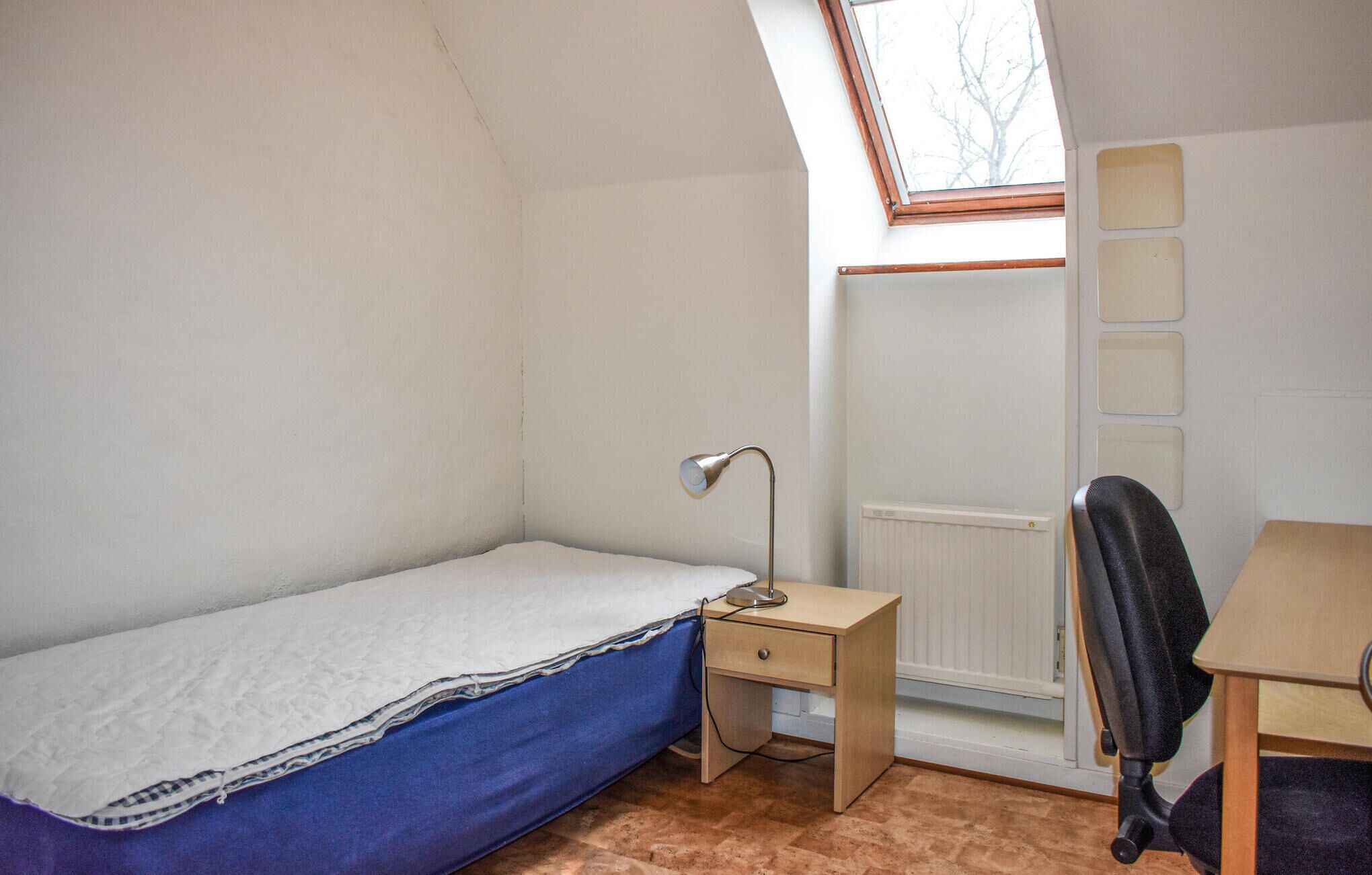 Une petite chambre avec un lit simple, un bureau et une chaise, un radiateur et une lucarne.