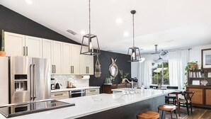 A modern kitchen with a large island, stainless steel appliances, pendant lighting, and a view of the outdoors. - Mid Century Home with Heated Pool + Spa (Bonita Springs)