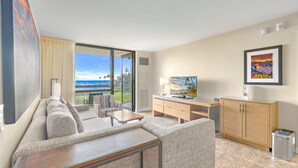A modern hotel room with a large sliding glass door leading to a balcony with a view of the ocean. There is a sofa, a small table, and a TV on a wooden cabinet.