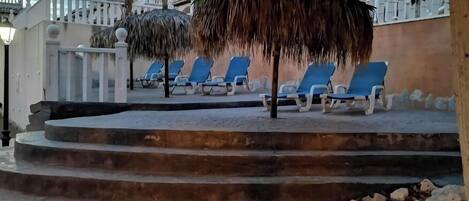 A poolside area with blue lounge chairs, a thatched umbrella, and a staircase leading down to a pebble beach.