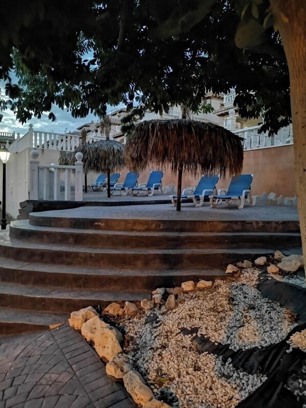 A poolside area with blue lounge chairs, a thatched umbrella, and a staircase leading down to a pebble beach.