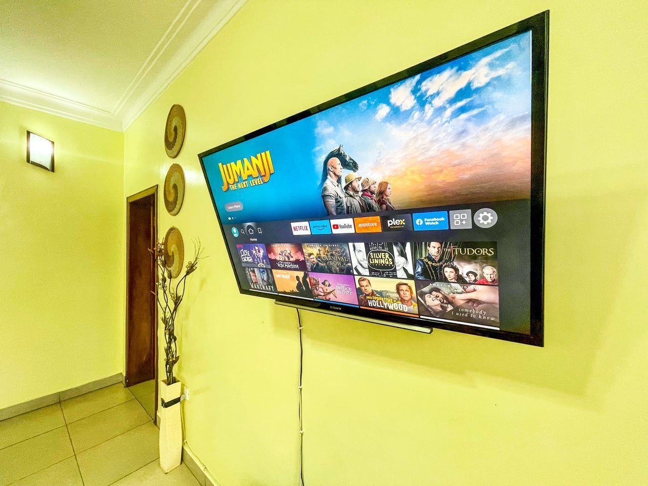 Un téléviseur à écran plat fixé sur un mur jaune, diffusant un film intitulé « Jumanji : Next Level », avec un ciel en arrière-plan.