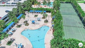 An aerial view of a resort with a swimming pool, sun loungers, and a tennis court.