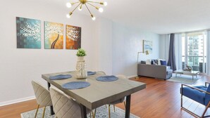 A modern living room with a dining area, featuring a dining table with blue placemats, a sofa, and a large window with a view of a building. - Komffy Apartment w/2 Queen Beds and panoramic view (Sunny Isles Beach)