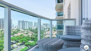 A balcony with wicker furniture, a glass railing, and a view of a cityscape with high-rise buildings and palm trees. - Komffy Apartment w/2 Queen Beds and panoramic view (Sunny Isles Beach)