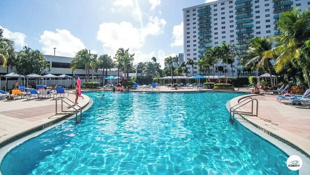A swimming pool with clear blue water, surrounded by lounge chairs and umbrellas, with a tall building in the background. - Komffy Apartment w/2 Queen Beds and panoramic view (Sunny Isles Beach)