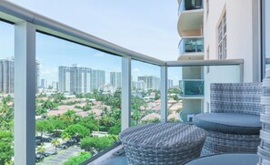 Un balcón con mobiliario de mimbre, barandilla de cristal y vistas a un paisaje urbano con rascacielos y palmeras.