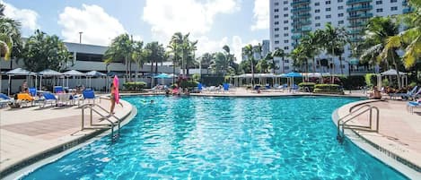 A swimming pool with clear blue water, surrounded by lounge chairs and umbrellas, with a tall building in the background.
