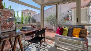 A sunlit room with a wooden desk, a chair, a colorful bench, and a large window overlooking a building and greenery. - Liberty 13 in Sassari (Sassari)