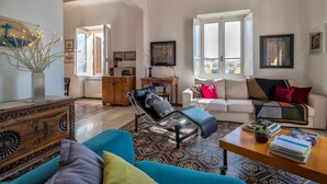 A spacious living room with a blue sofa, a black armchair, and a white sofa with colorful cushions. There is a wooden cabinet, a small table with books and a cactus, and a large window with shutters. - Liberty 13 in Sassari (Sassari)
