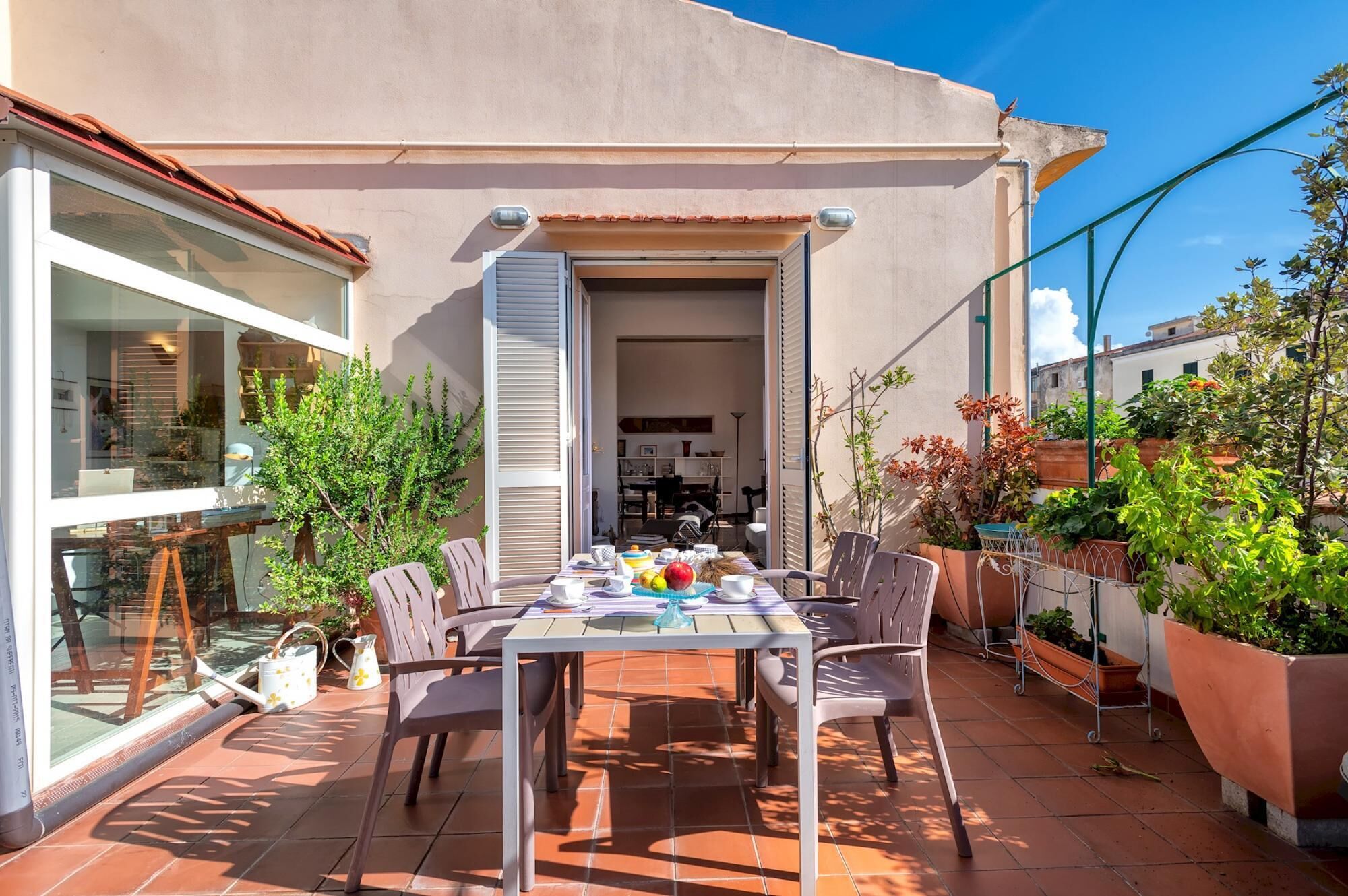 Area pranzo all'aperto con tavolo apparecchiato per quattro persone, circondato da sedie, su un patio piastrellato.