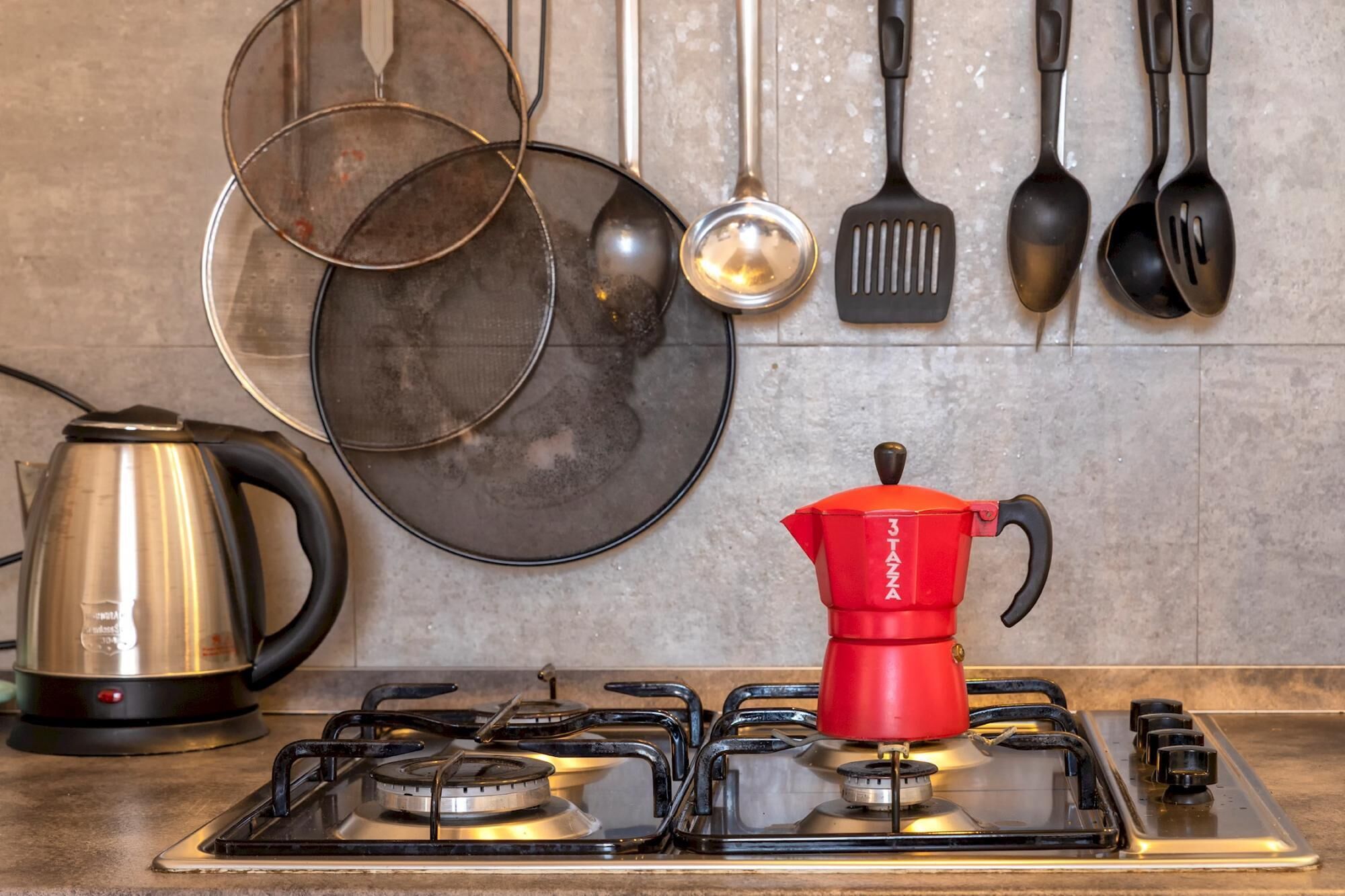 Une cuisine équipée d’une cuisinière à gaz, d’une cafetière rouge, d’une bouilloire en acier inoxydable et de divers ustensiles de cuisine accrochés au mur.