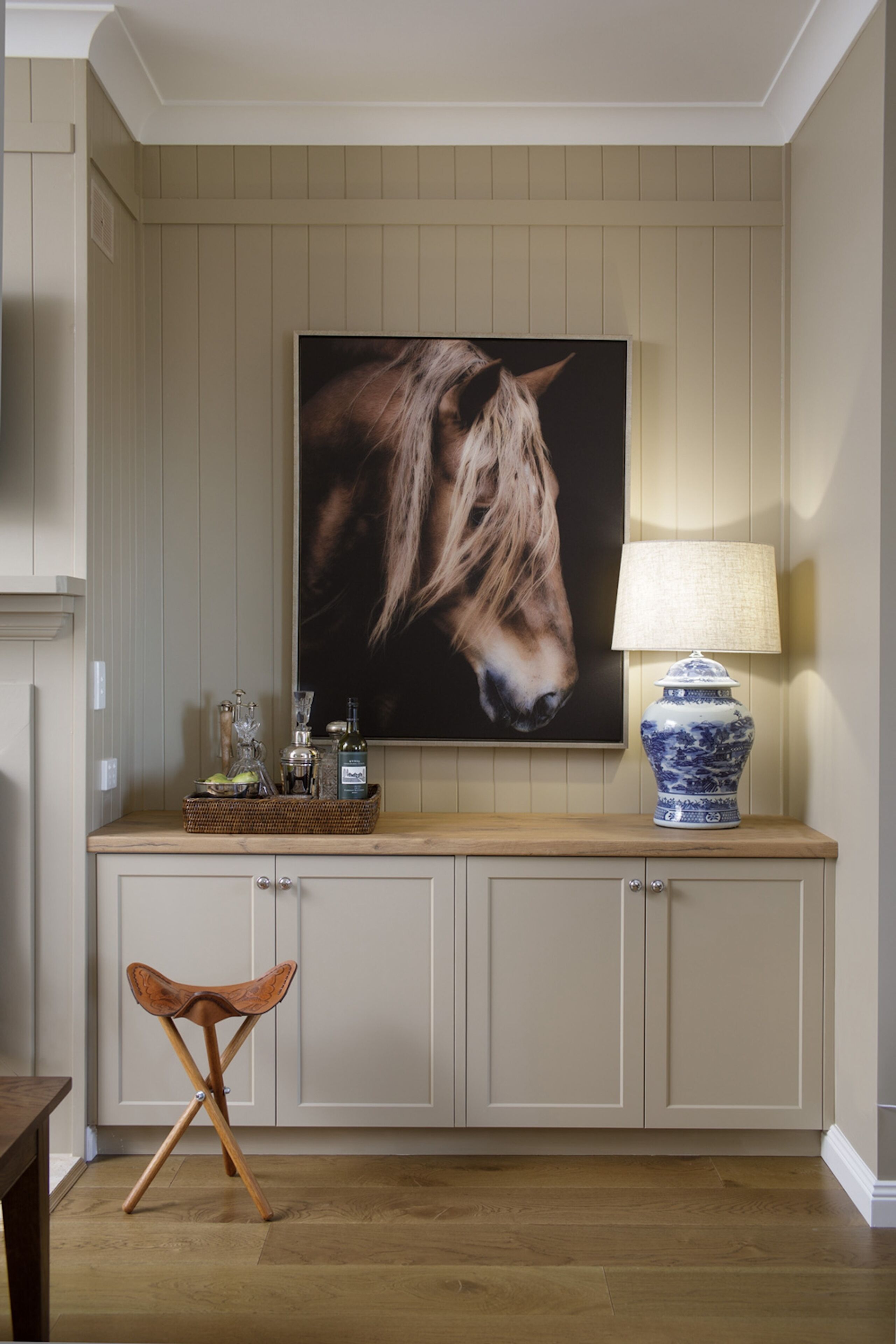 A wooden cabinet with a mounted painting of a horse's head, a lamp, and a wicker basket with bottles.