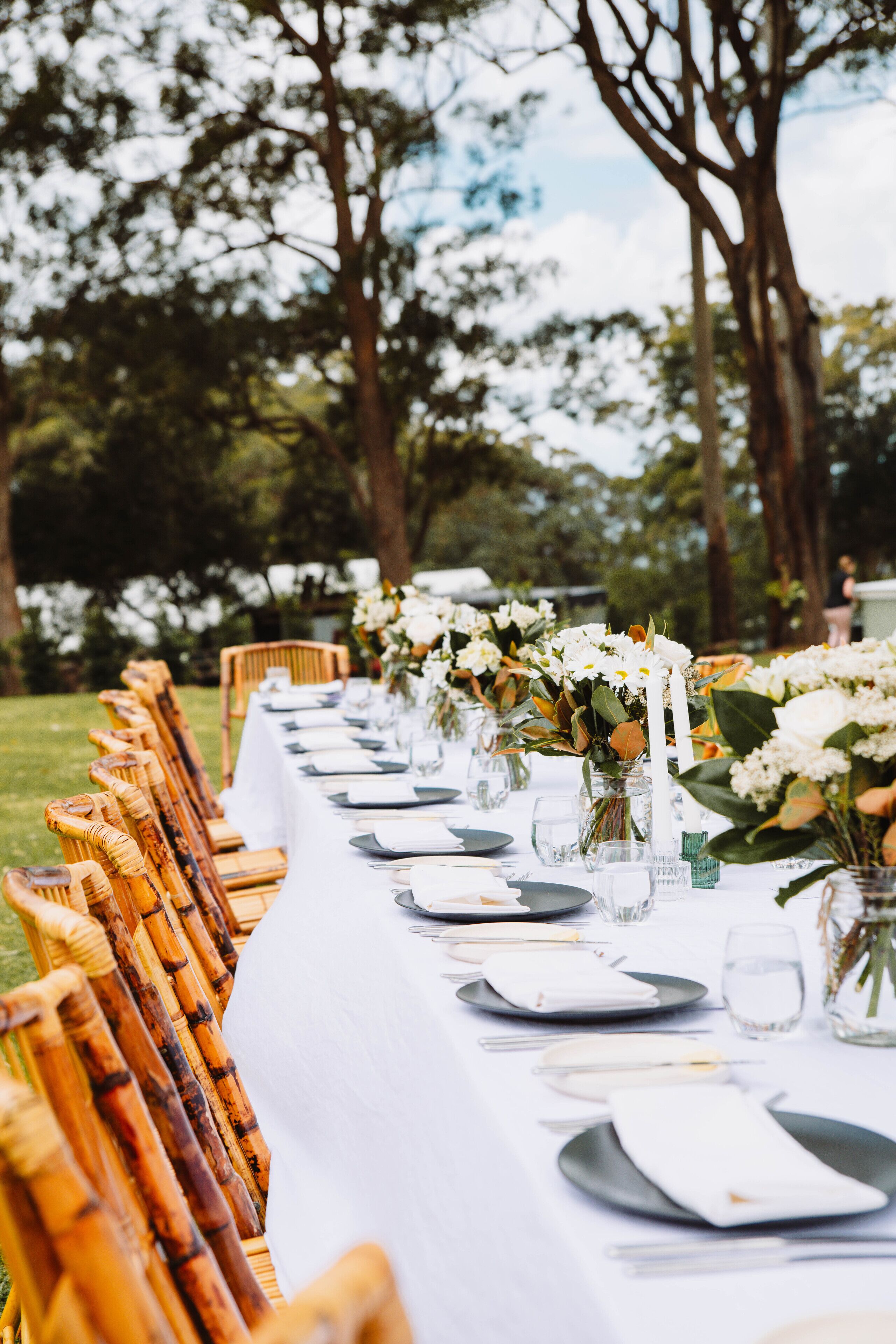 A long table set outdoors with chairs, white tablecloth, and floral centerpieces.