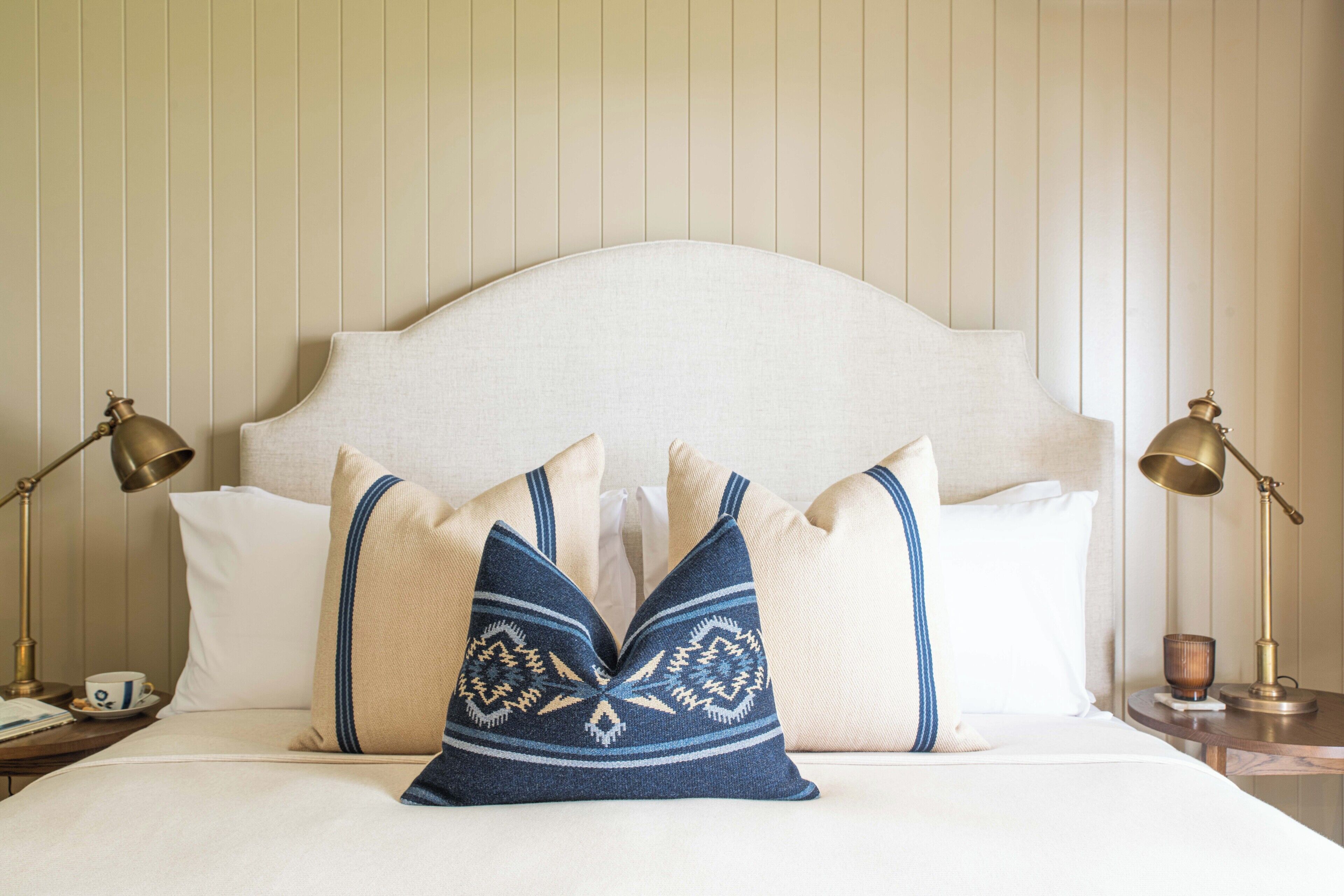 Cama bem arrumada com cabeceira branca, duas luminárias de mesa e travesseiros decorativos.