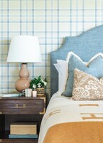 Una cama bien hecha con cabecera azul, una lámpara de noche, una planta pequeña y un libro titulado 