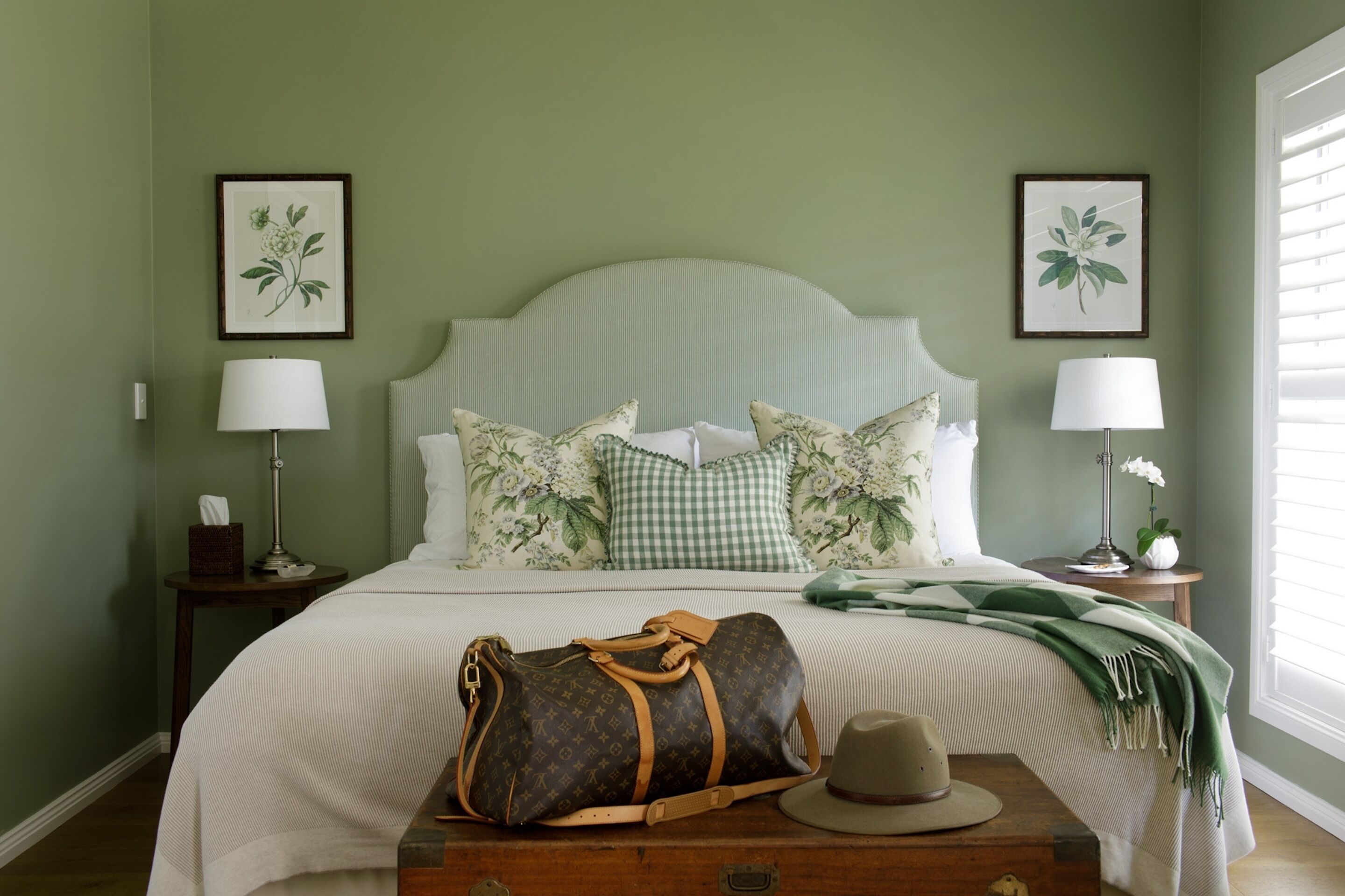 A neatly made bed with floral pillows, a green blanket, and a Louis Vuitton bag on a wooden chest.