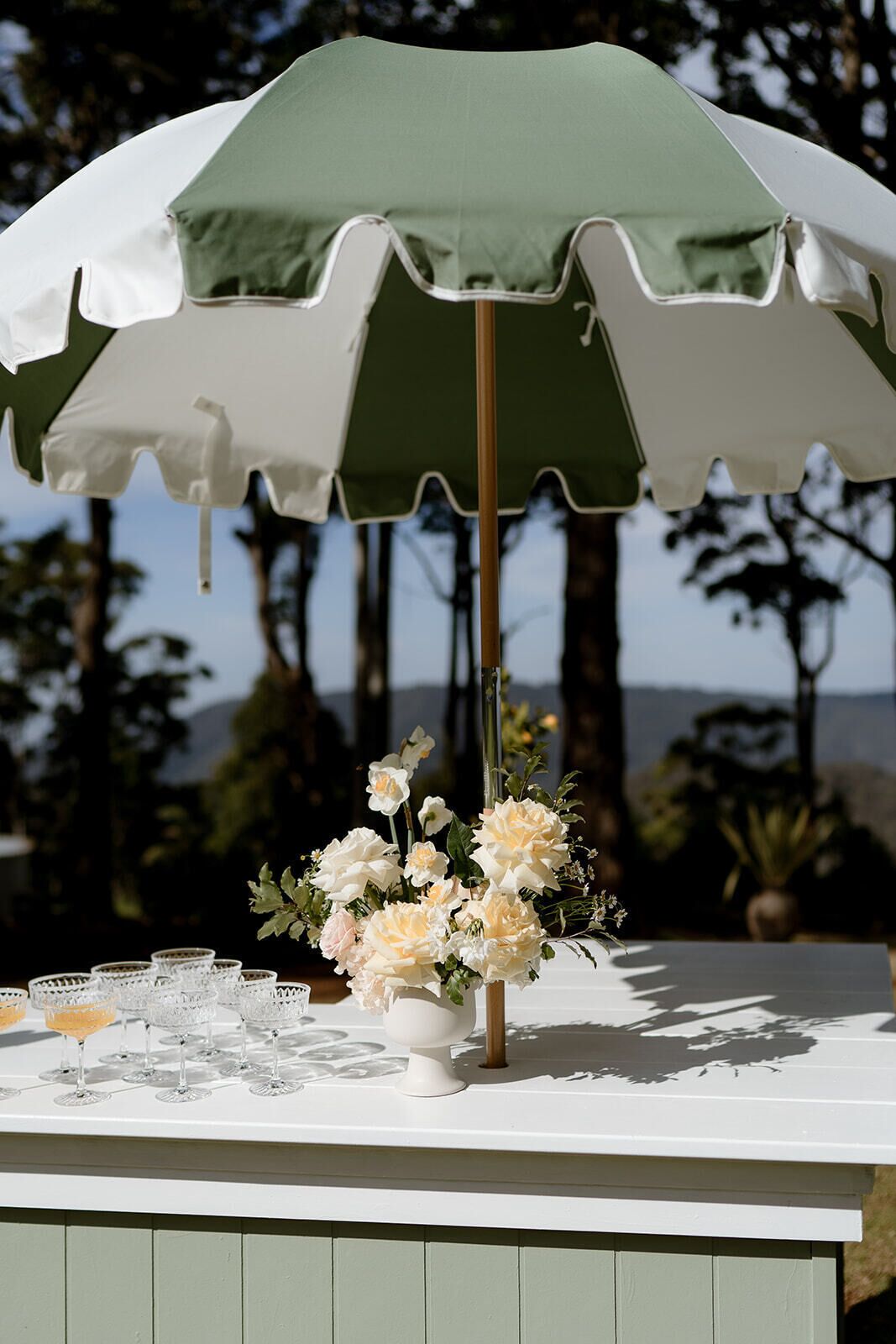 A table with a green and white umbrella, a vase with flowers, and crystal glasses.