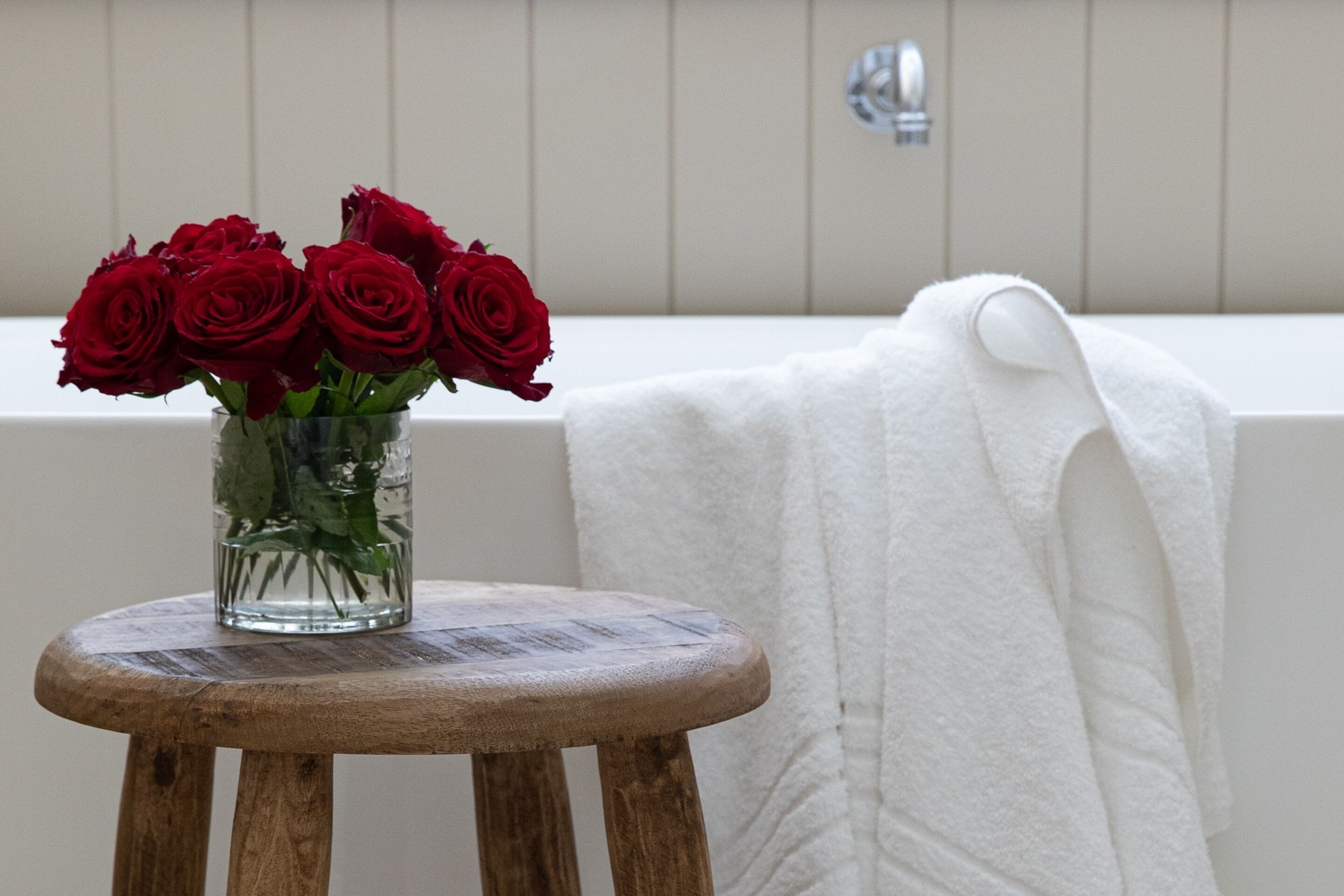 A bathroom with a bathtub, a white towel, and a wooden stool with a vase of red roses.