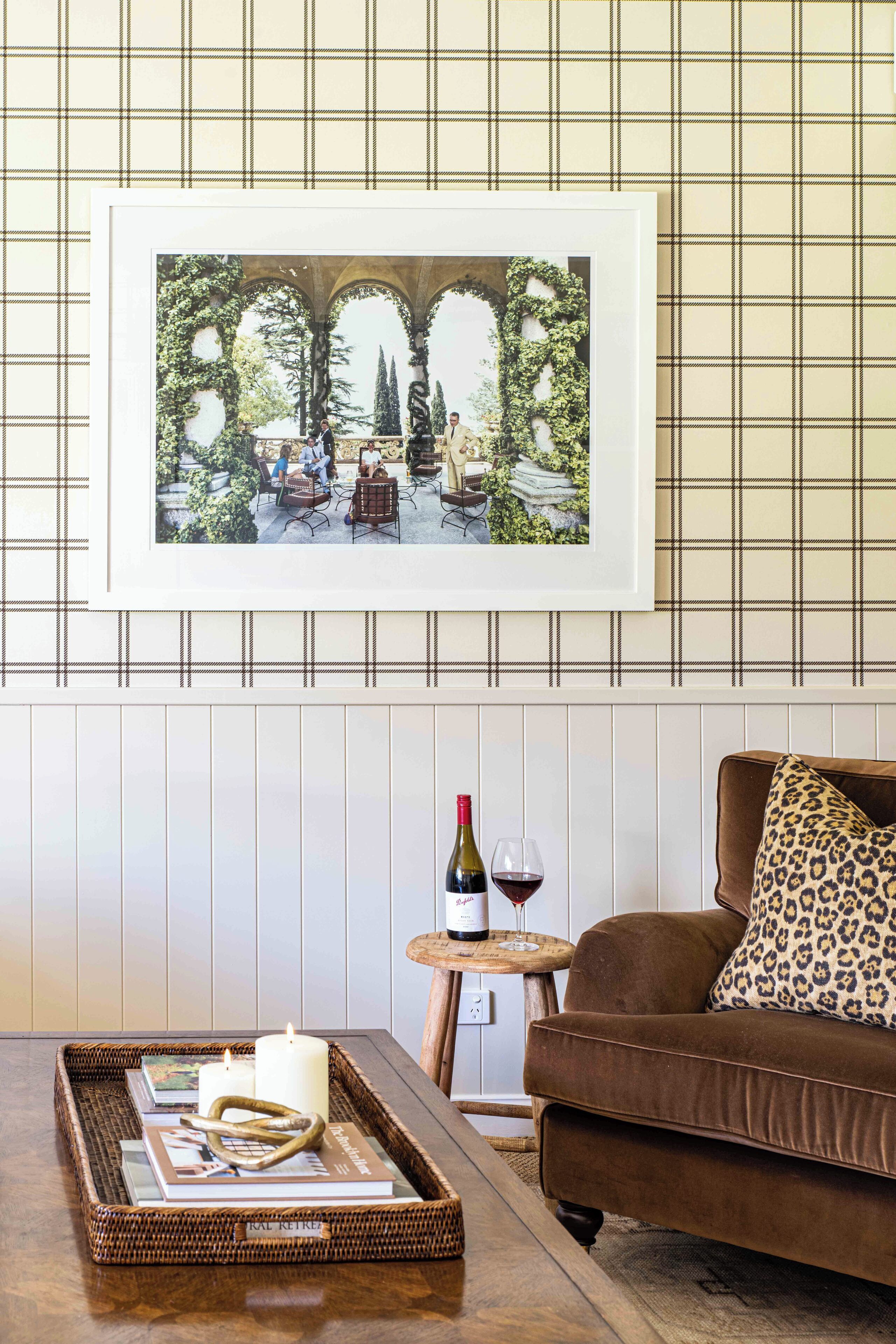 A cozy living room with a brown sofa, a small round table with a wine bottle and glass, and a wicker tray with books and a candle.