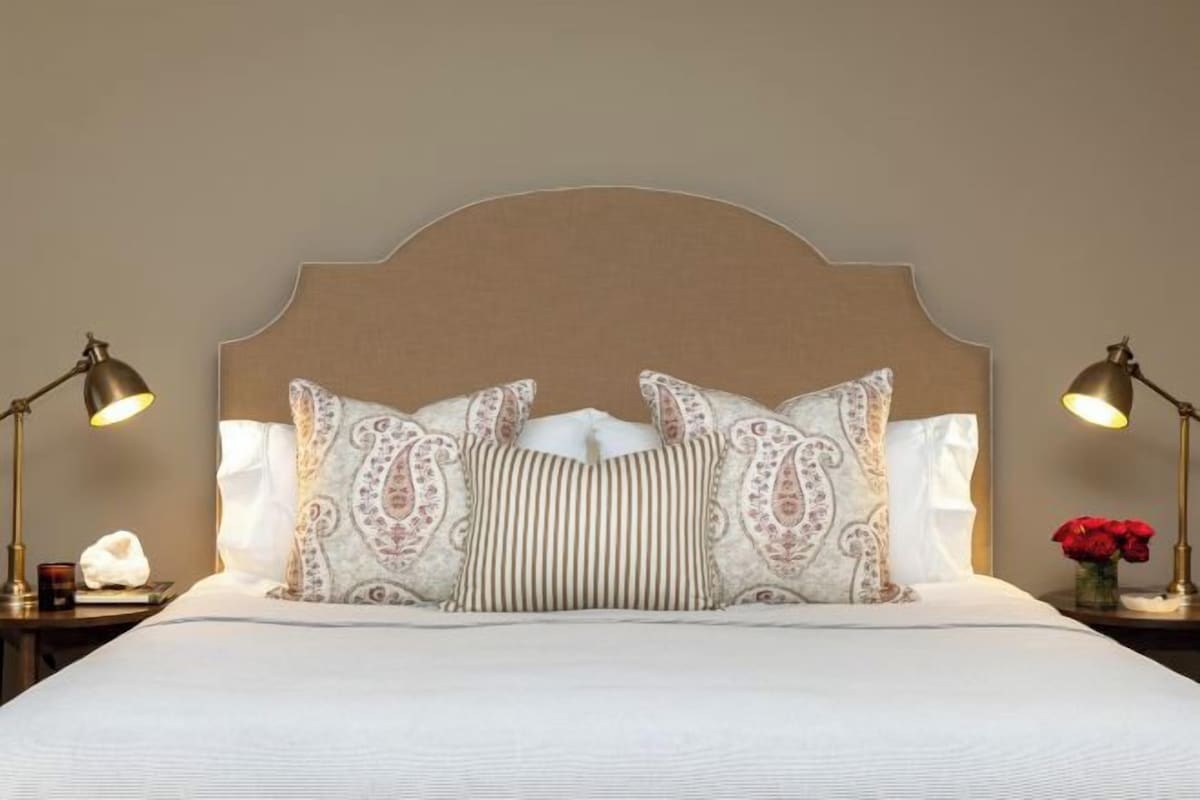 A neatly made bed with patterned pillows, a headboard, bedside lamps, and a vase with flowers.