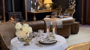 Un comedor acogedor con una mesa redonda dispuesta para cuatro personas, rodeada de sillas de mimbre. Se observa una araña colgante y una chimenea al fondo.