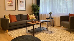 Un salon moderne avec un canapé gris, une table basse et une table d’appoint. Deux tableaux encadrés ornent le mur, ainsi qu’une plante en pot et une fenêtre agrémentée de rideaux légers.