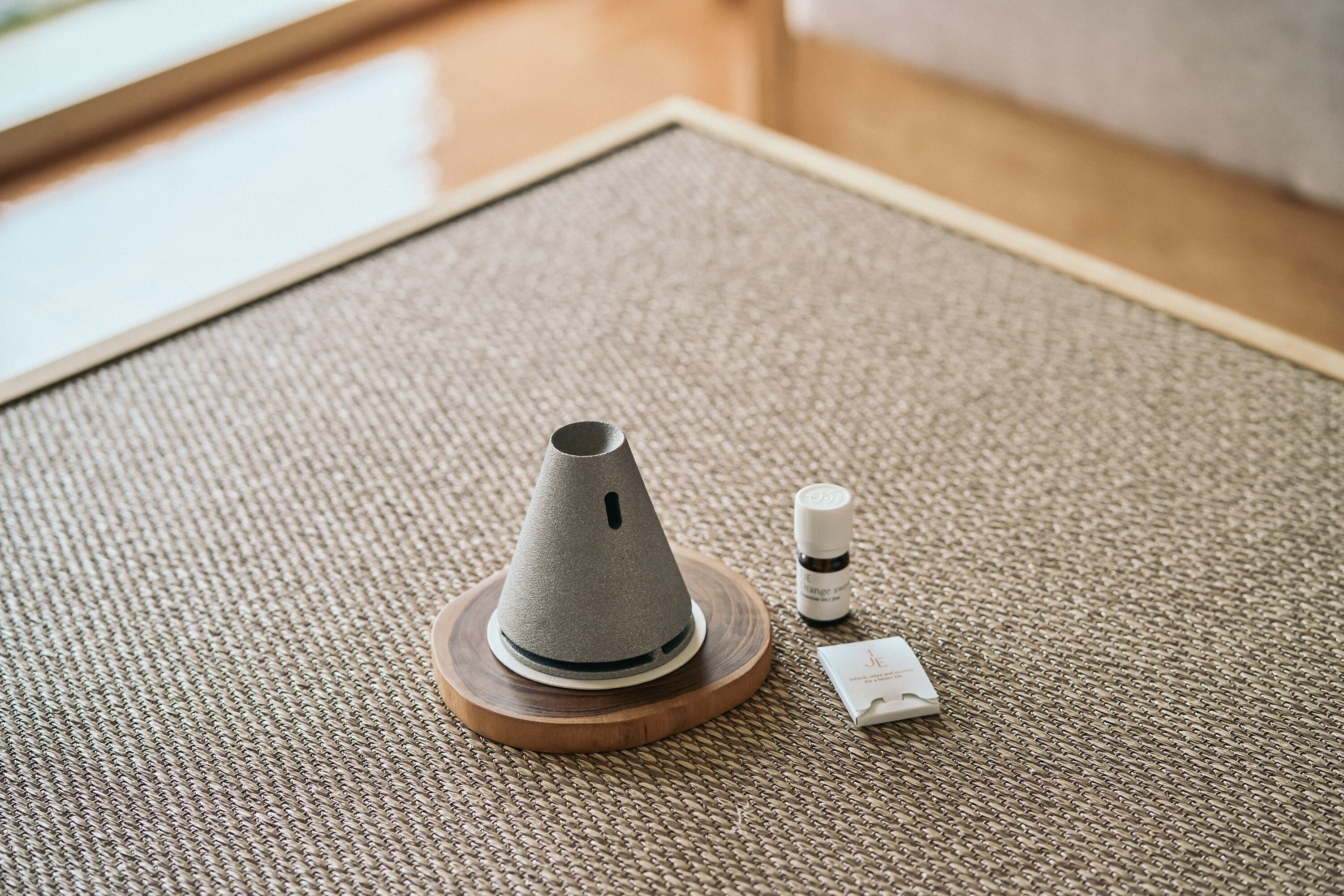 Une petite table ronde en bois, sur laquelle est posé un tapis tissé. Sur la table, on trouve un objet gris en forme de cône, une petite bouteille et une boîte blanche.