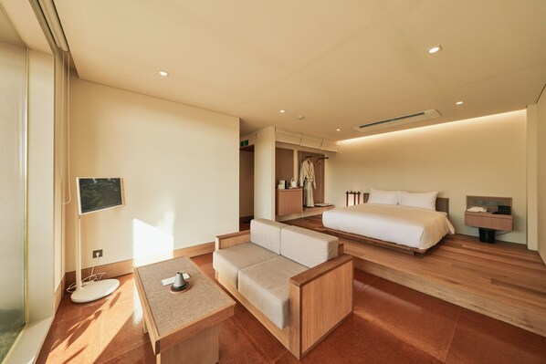 A modern hotel room with a wooden floor, a sofa, a bed, a bedside table, and a small desk with a lamp.