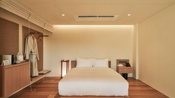 A modern hotel room with a large bed, wooden flooring, a built-in desk, a robe rack, and a small bedside table with a phone and a lamp.