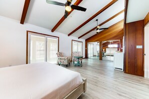 Een ruime slaapkamer met een groot bed, een plafondventilator en uitzicht op de keuken en woonkamer door een glazen deur.
